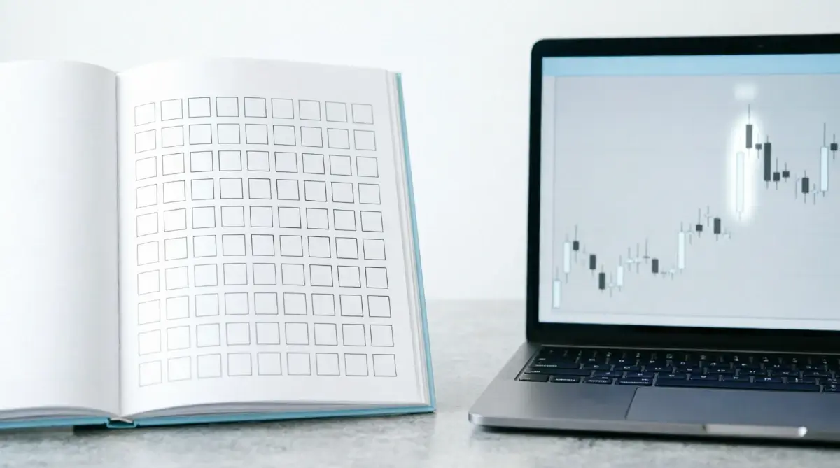 Open journal with grid of empty checkboxes next to laptop showing historical price chart pattern